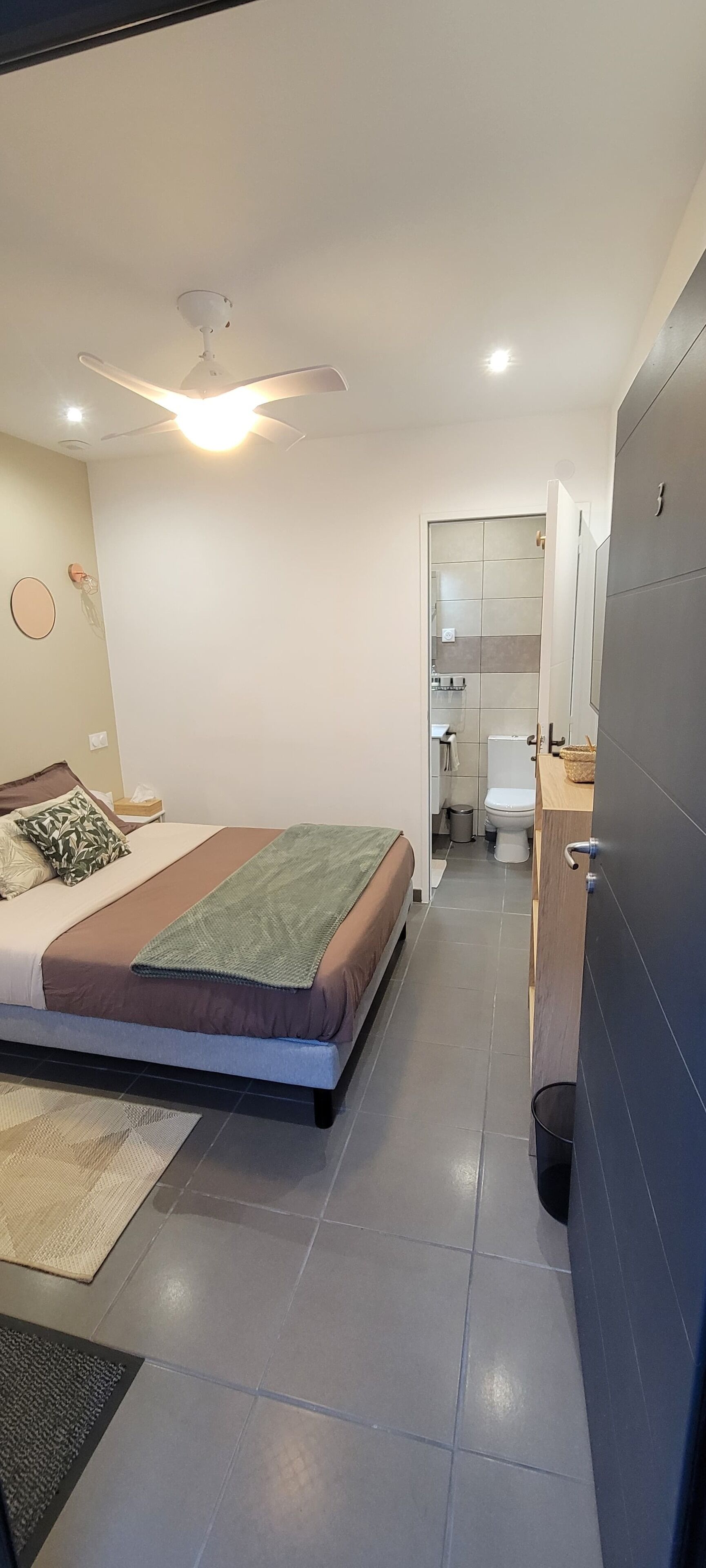 Ein modernes Schlafzimmer mit Bett, Deckenventilator, Bad mit Toilette und Waschbecken sowie einem großen Spiegel.