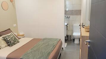 Ein modernes Schlafzimmer mit Bett, Deckenventilator, Bad mit Toilette und Waschbecken sowie einem großen Spiegel.
