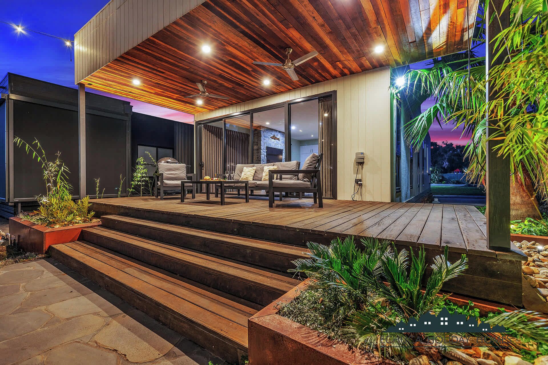 A modern outdoor patio with a wooden deck, a seating area with a sofa and chairs, and a staircase leading to the entrance.