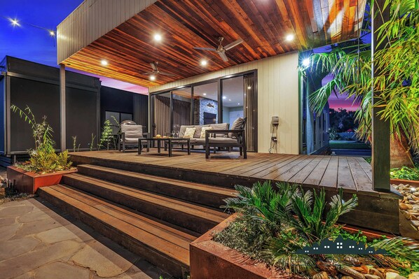 A modern outdoor patio with a wooden deck, a seating area with a sofa and chairs, and a staircase leading to the entrance.