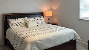 A bedroom with a bed, a ceiling fan, a nightstand with a lamp, and a window with blinds.