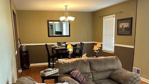 A living room with a sectional sofa, a dining table with chairs, a chandelier, and a large window with blinds.