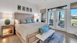 A bedroom with a large bed, a nightstand with a lamp, a bench, and a view of the outdoors through a sliding door. - Lakefront 402, Upgraded 3-bd, 3-bath lakefront villa just steps to the beach! (Miramar Beach)