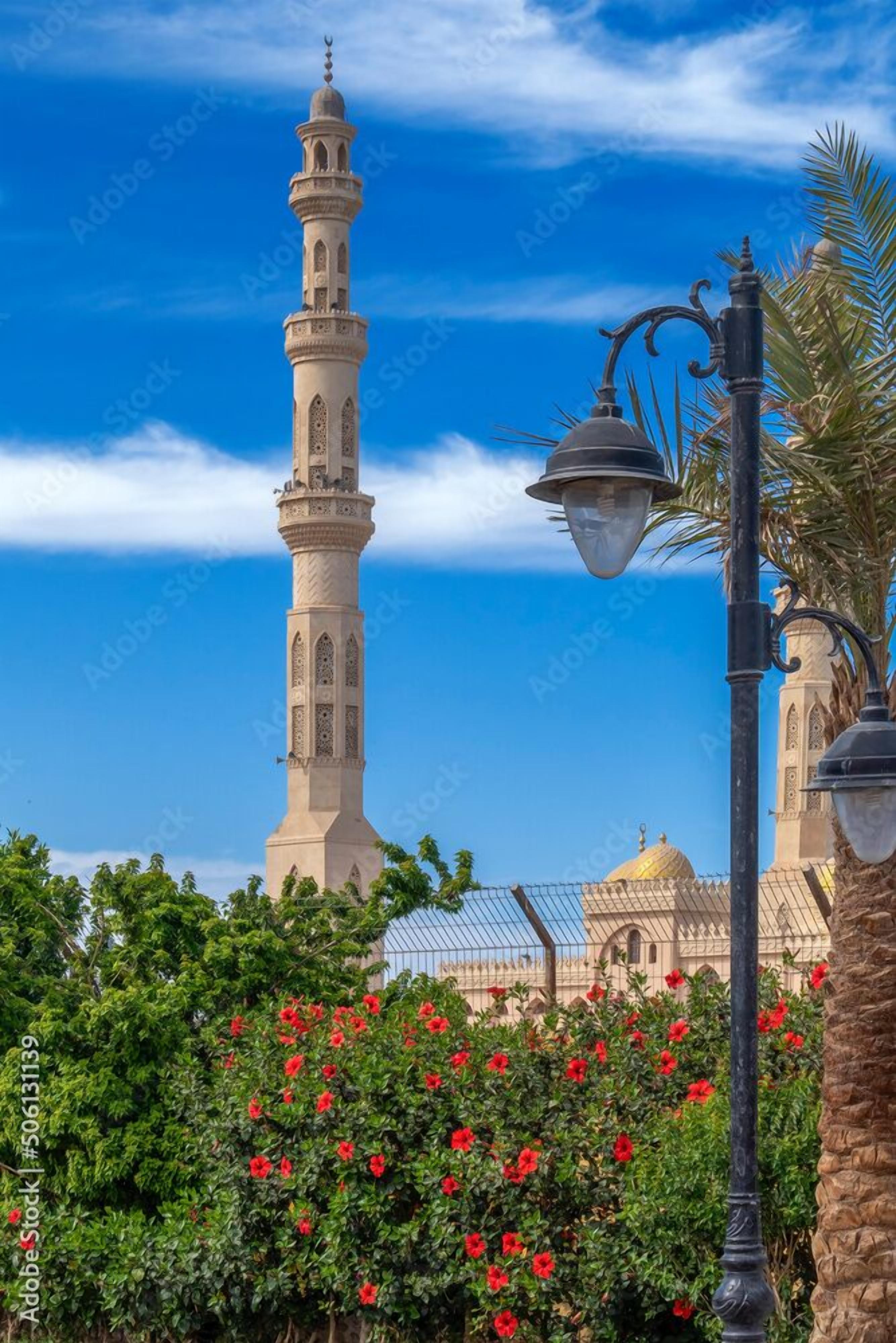A tall minaret with intricate designs, a mosque with a golden dome, and a lush garden with red flowers.