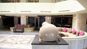 Eine großzügige Lobby mit einer zentralen Sitzecke, die ein großes Steinskulptur, gepolsterte Sitzmöbel mit rosa und beige Kissen sowie einen Steinfußboden mit geometrischem Muster zeigt.