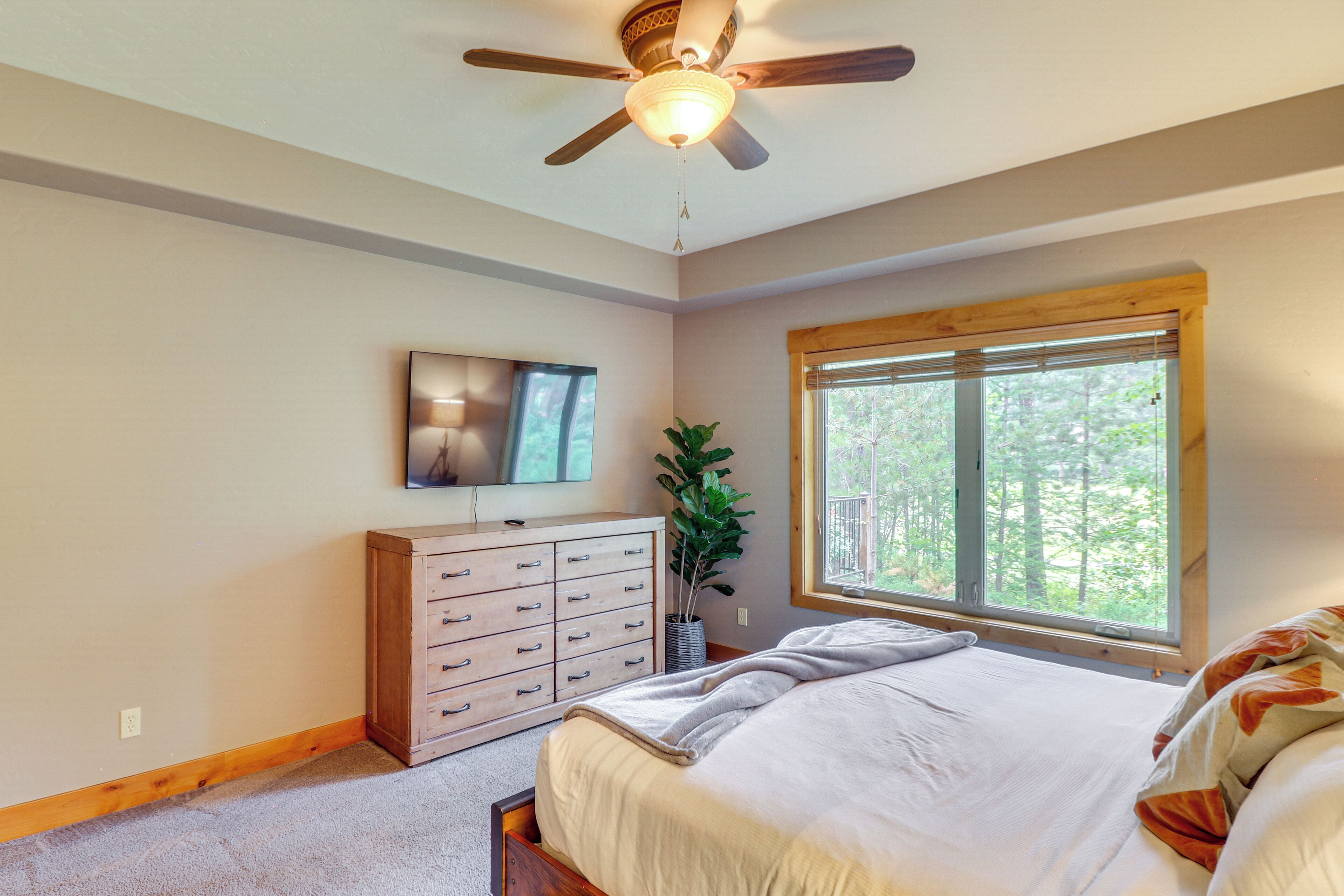 A bedroom with a bed, a wooden dresser, a ceiling fan, a window with a view of trees, and a television on the wall.