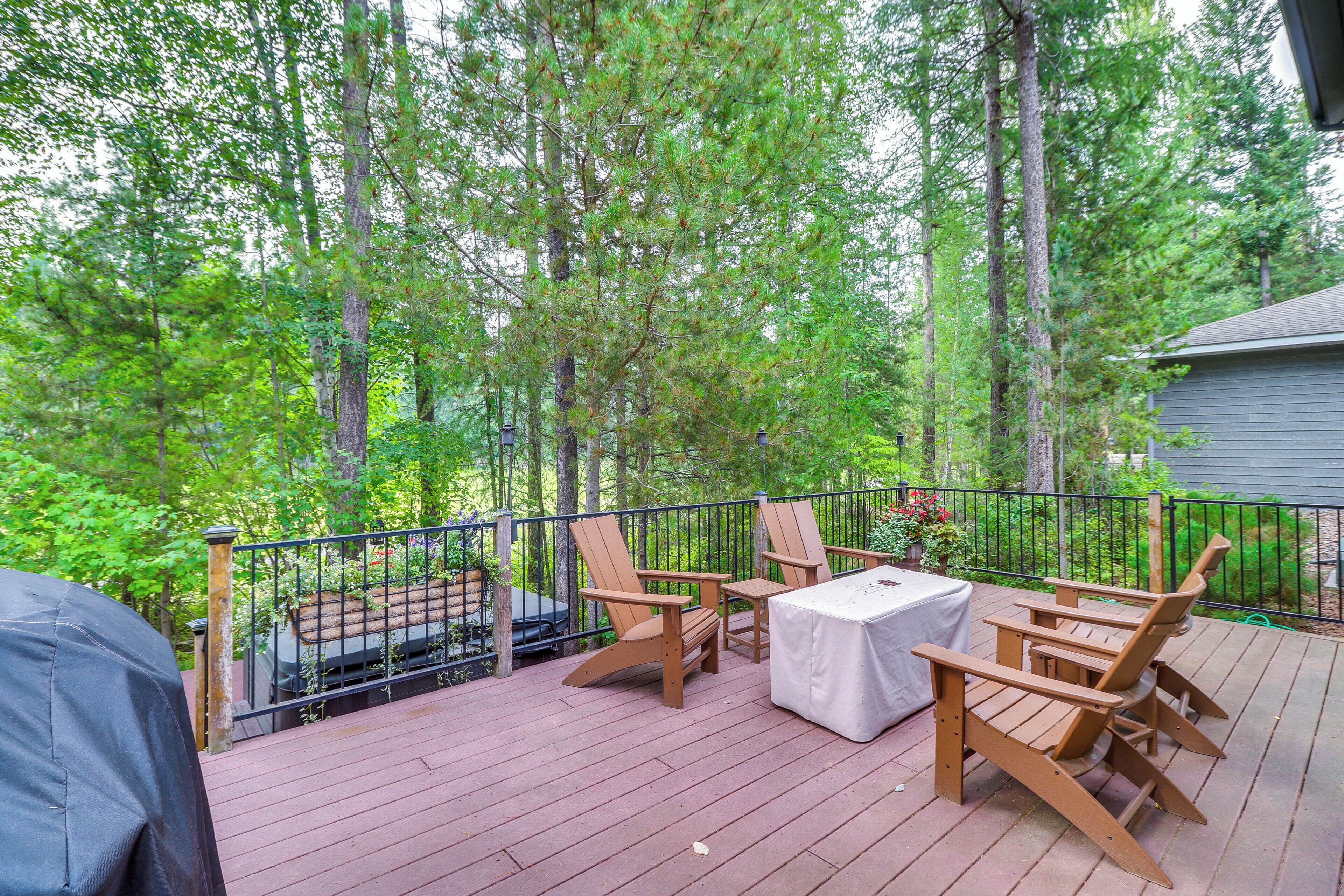 A wooden deck with Adirondack chairs and a table, surrounded by a metal railing and dense trees.
