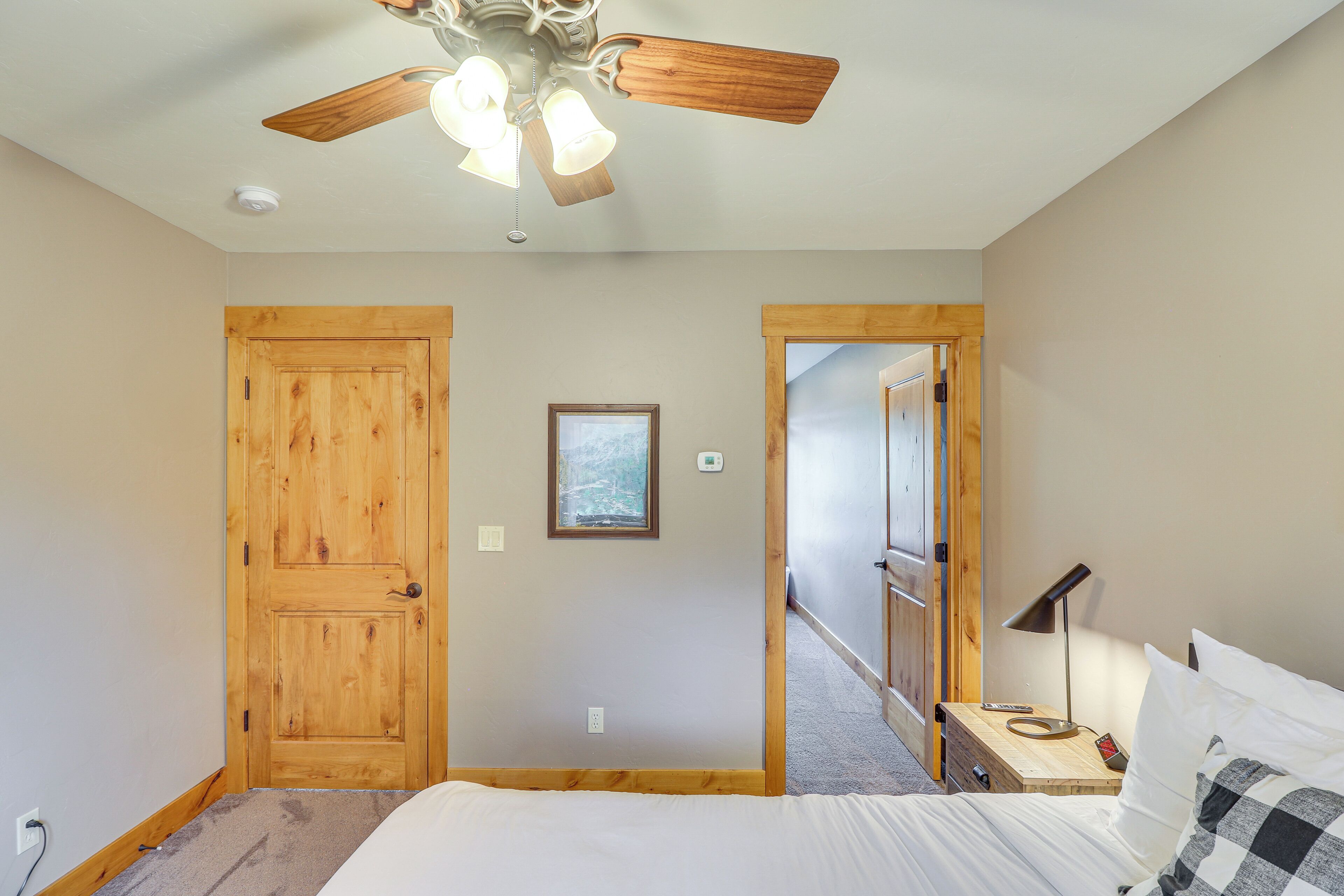 A bedroom with a wooden door, a ceiling fan, a bedside table with a lamp, and a bed.
