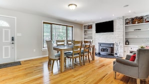 A dining area with a wooden table and chairs, a fireplace, and a TV mounted on a white brick wall. - Hot Tub + Fire Pit: Pocono Summit Home! (Pocono Summit)