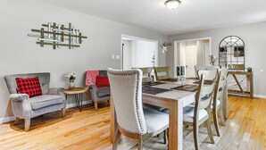 A dining area with a wooden table and chairs, a grey sofa with a red pillow, and a wall-mounted decorative piece. - Hot Tub + Fire Pit: Pocono Summit Home! (Pocono Summit)