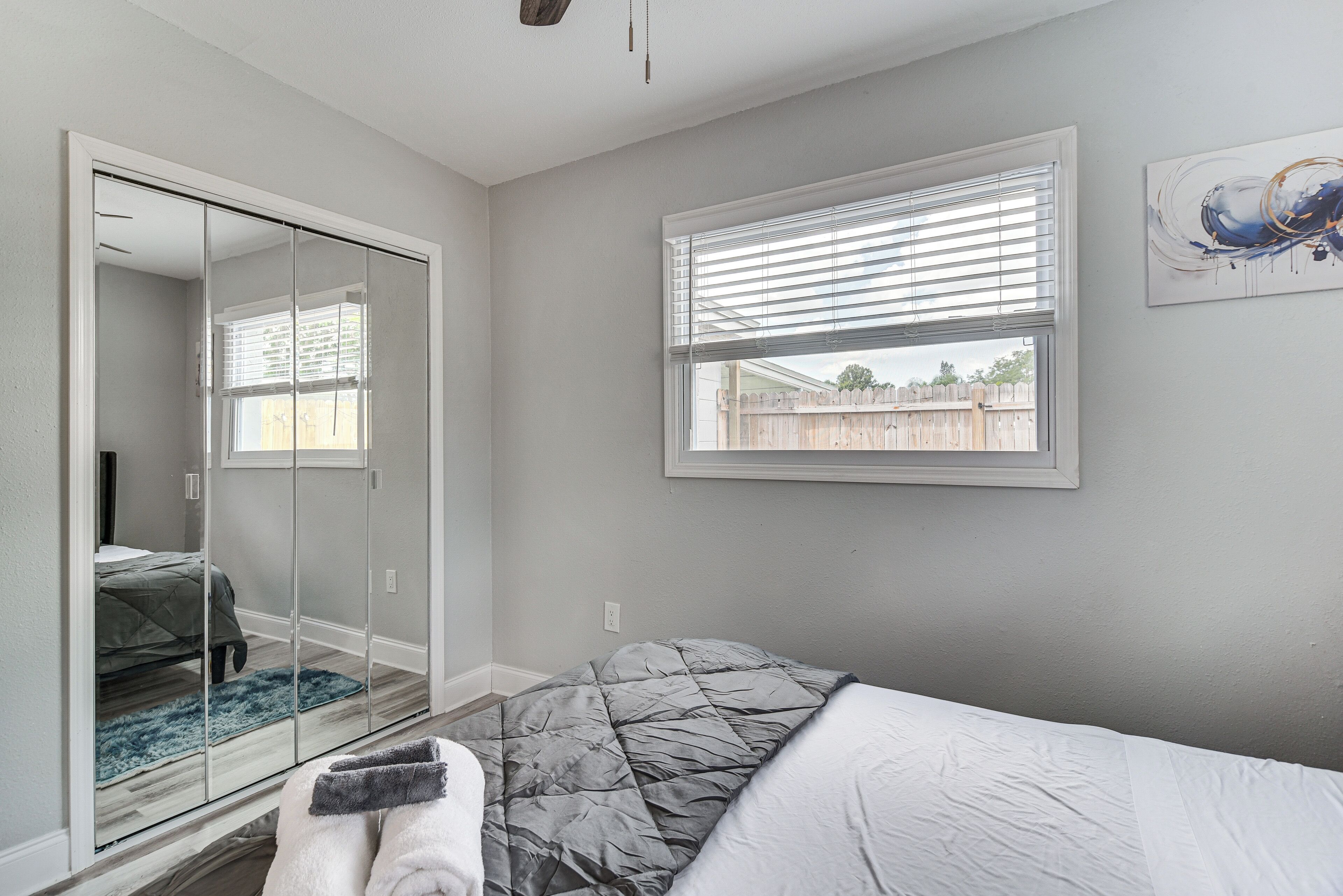 Une chambre à coucher avec un lit, un grand miroir, une fenêtre avec des stores et une vue sur une clôture.