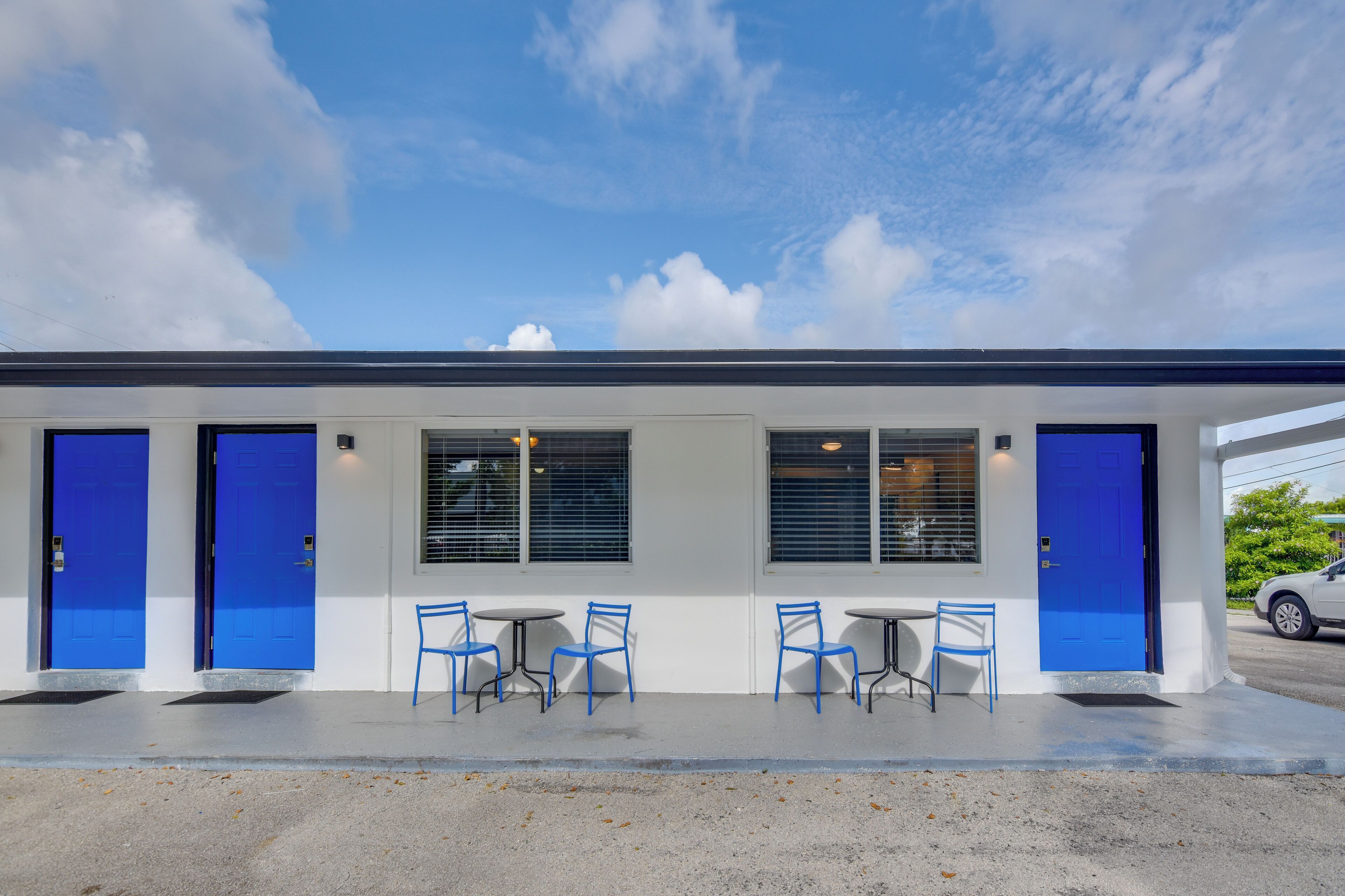 Un bâtiment avec deux portes bleues, un espace extérieur abrité avec des chaises bleues, et une façade blanche.