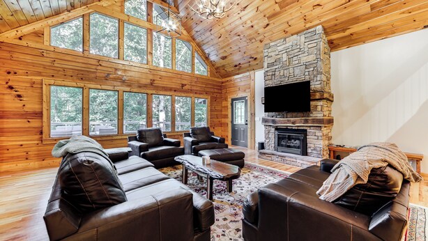 A log cabin interior with a stone fireplace, leather sofas, and large windows. - Luxe Smoky Mountains Cabin w/ Hot Tub & Views! (Waynesville)