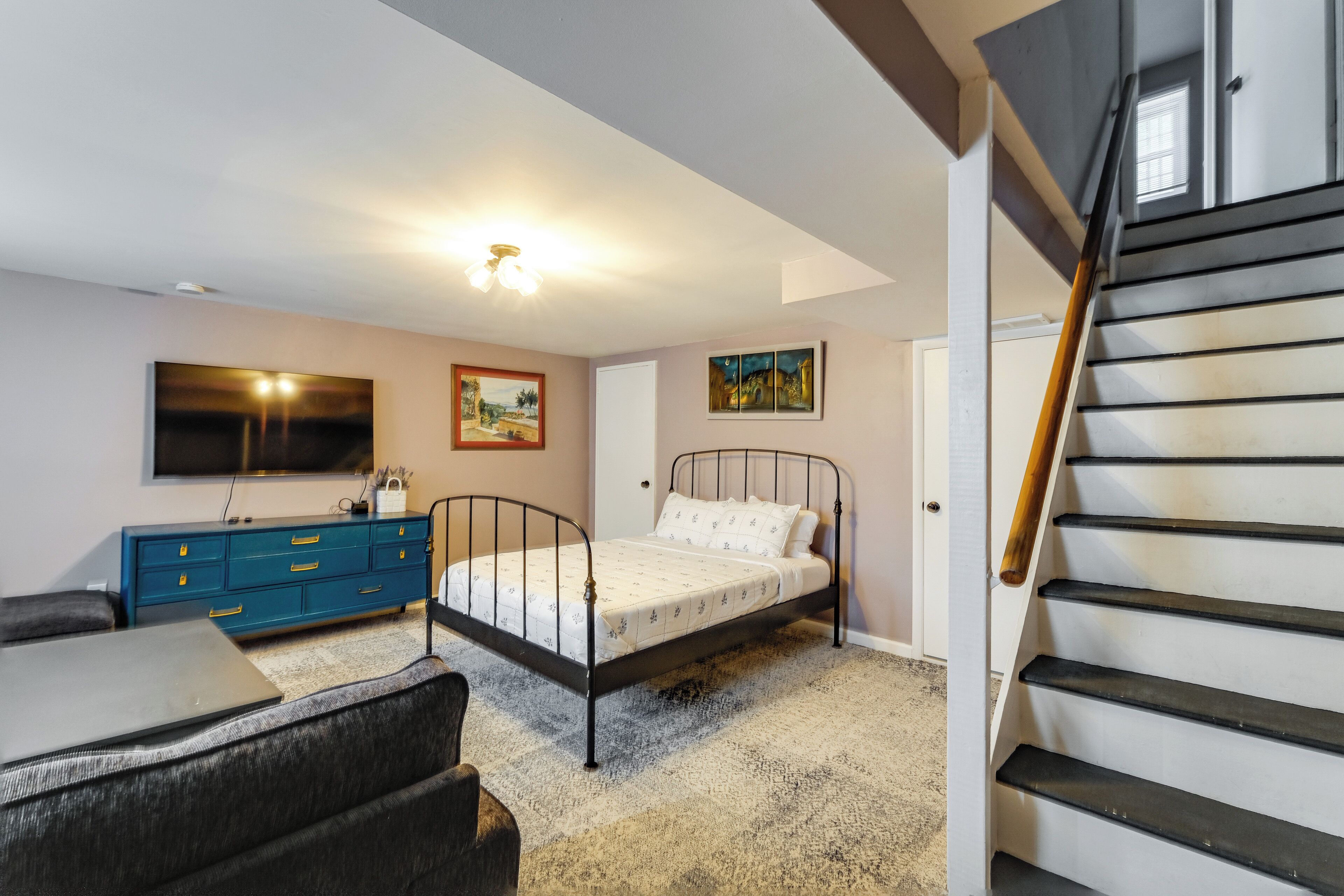 A bedroom with a bed, a dresser, a TV, a painting, and a staircase.