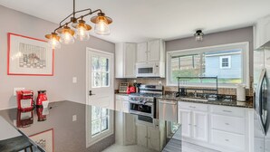 Une cuisine moderne avec des armoires blanches, un réfrigérateur en acier inoxydable, un four à micro-ondes et une cuisinière à gaz. On y trouve un îlot central en verre, sur lequel sont posés une cafetière rouge et une bouilloire.