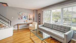 Un salon moderne avec un canapé gris, une table basse en verre, un fauteuil blanc et un tableau abstrait aux couleurs vives accroché au mur.