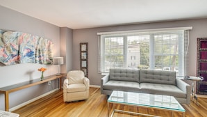 Un salon avec un canapé gris, un fauteuil beige, une table basse en verre, un banc en bois et un grand tableau abstrait accroché au mur.