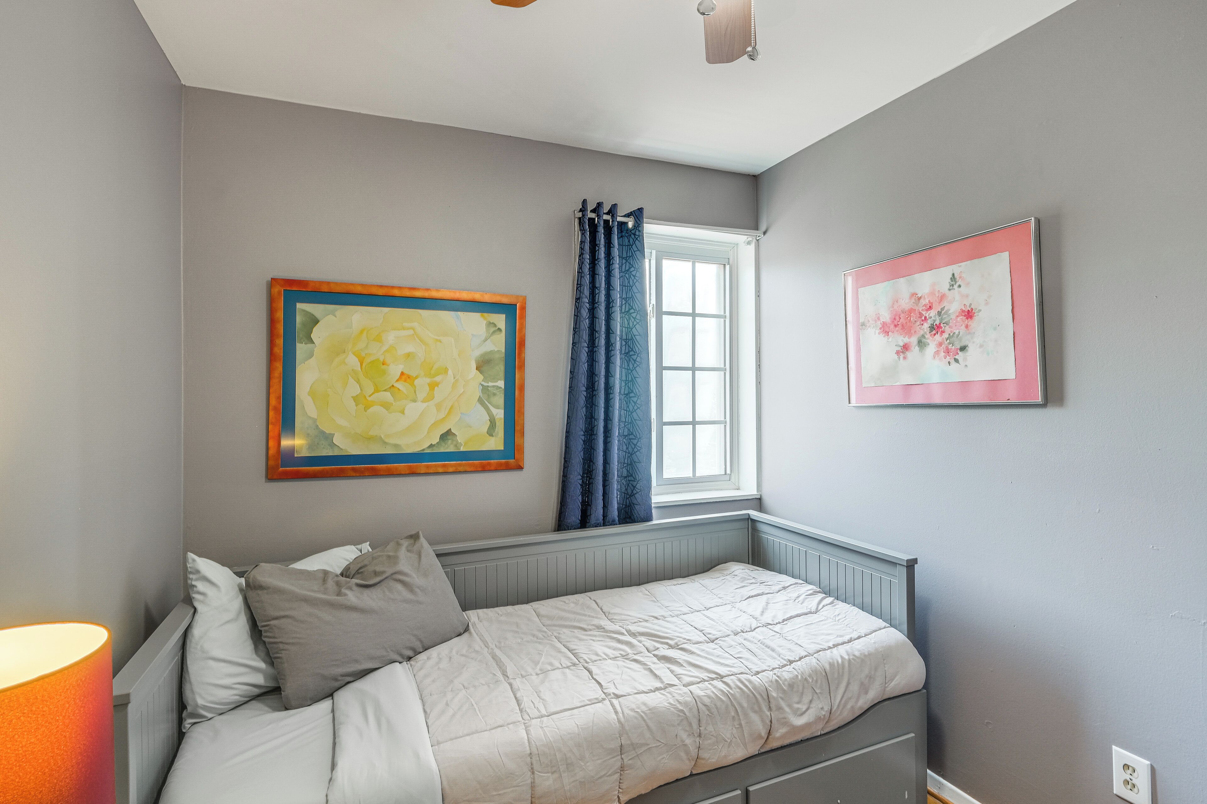 Ein Schlafzimmer mit einem Einzelbett, einem Fenster mit Vorhängen und zwei gerahmten Kunstwerken an den Wänden.