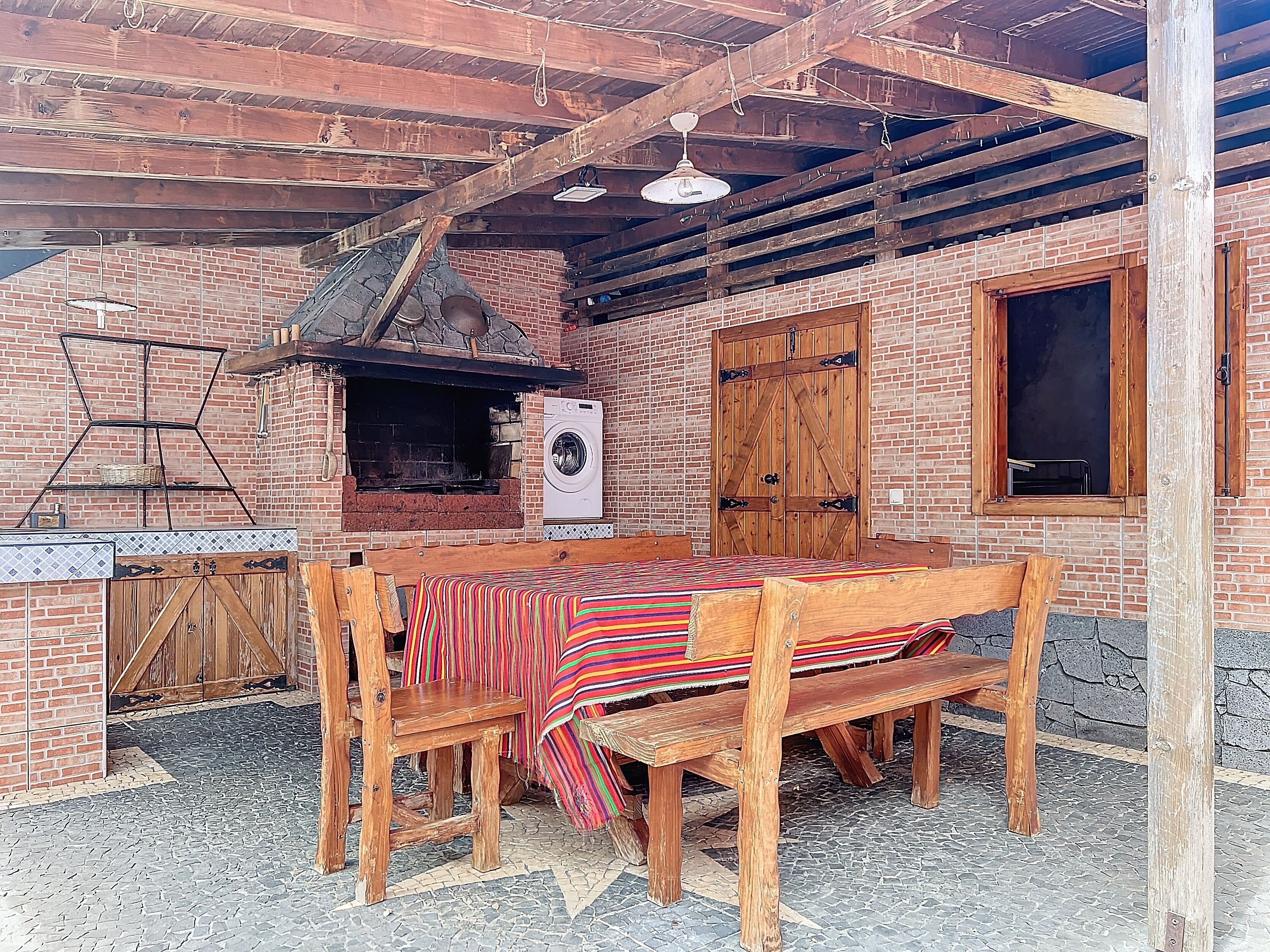 Une cuisine extérieure rustique avec un mur en briques, des poutres en bois, un luminaire suspendu, un lave-linge et une table en bois recouverte d’une nappe colorée.