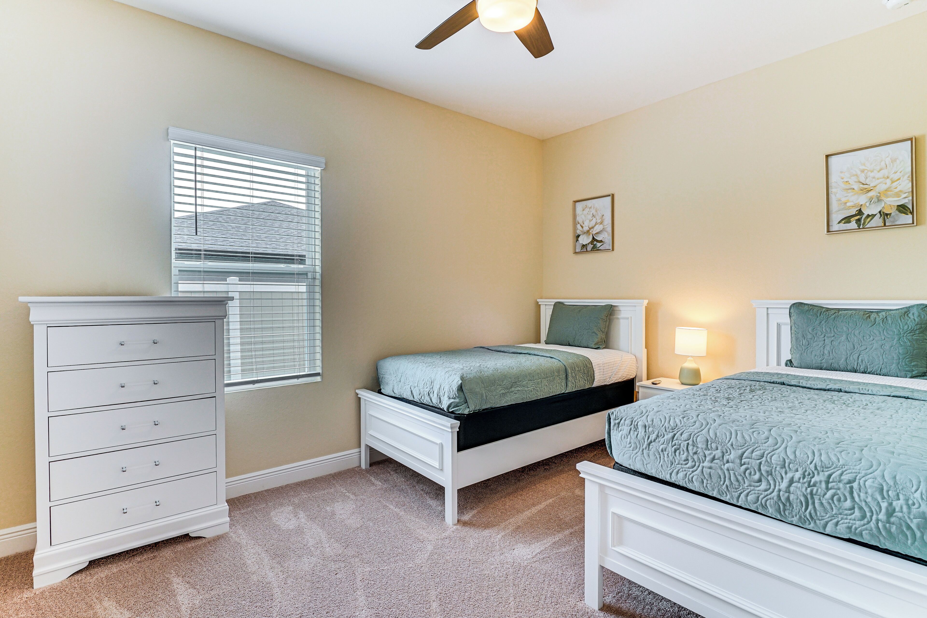 A bedroom with a white dresser, a bed with a green bedspread, a nightstand with a lamp, and a ceiling fan.