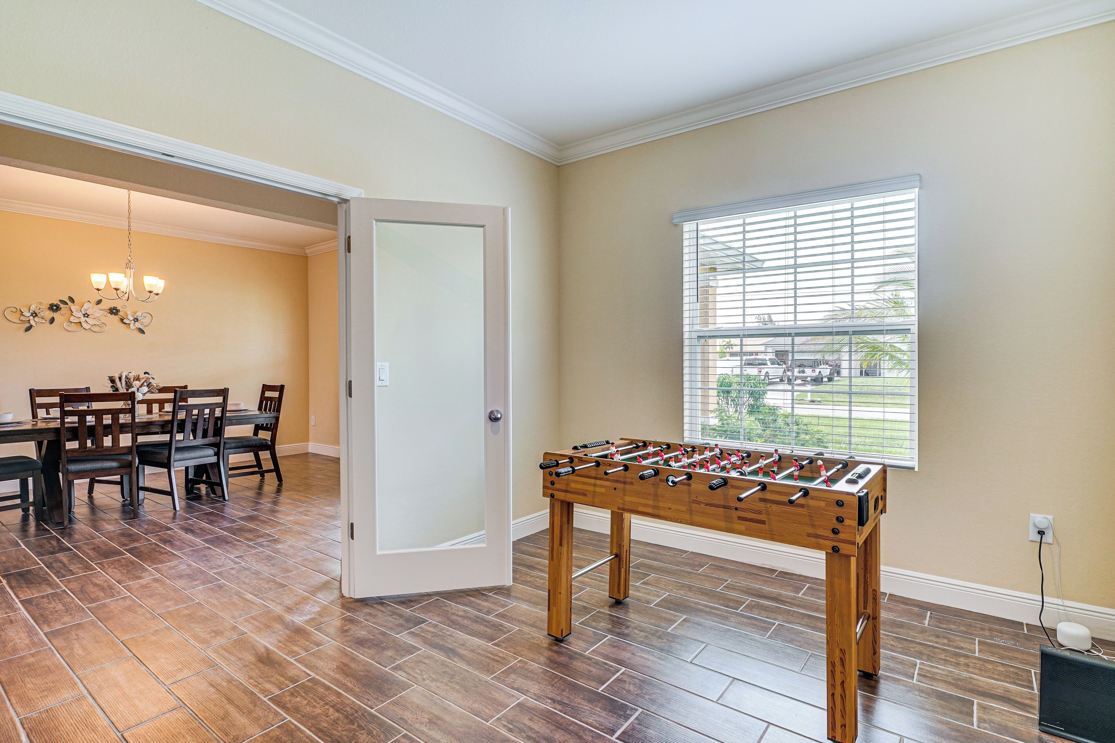 A room with a foosball table, a dining area with a chandelier, and a window with blinds.