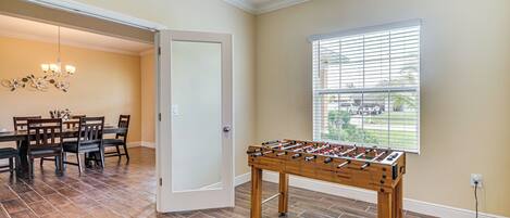 A room with a foosball table, a dining area with a chandelier, and a window with blinds.