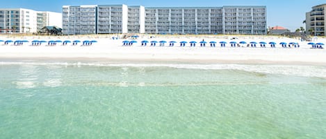 Un hotel frente a la playa con varios pisos, una playa arenosa y sombrillas azules.
