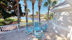 Een houten terras met blauwe Adirondack-stoelen, een witte reling en uitzicht op een meer met palmbomen.