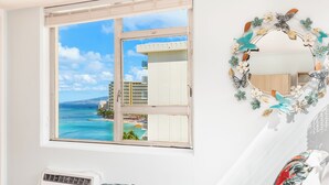 Une chambre dotée d’une grande fenêtre donnant sur l’océan et les bâtiments, d’un climatiseur blanc et d’une couvre-lit à motifs floraux.