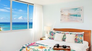 Une chambre d’hôtel dotée d’une grande fenêtre donnant sur l’océan, d’un lit avec une literie à motifs floraux et d’un tableau représentant une scène de plage.