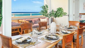 Un espace repas offrant une vue sur l’océan, avec des meubles en bois et un arrangement floral en guise de décoration centrale.