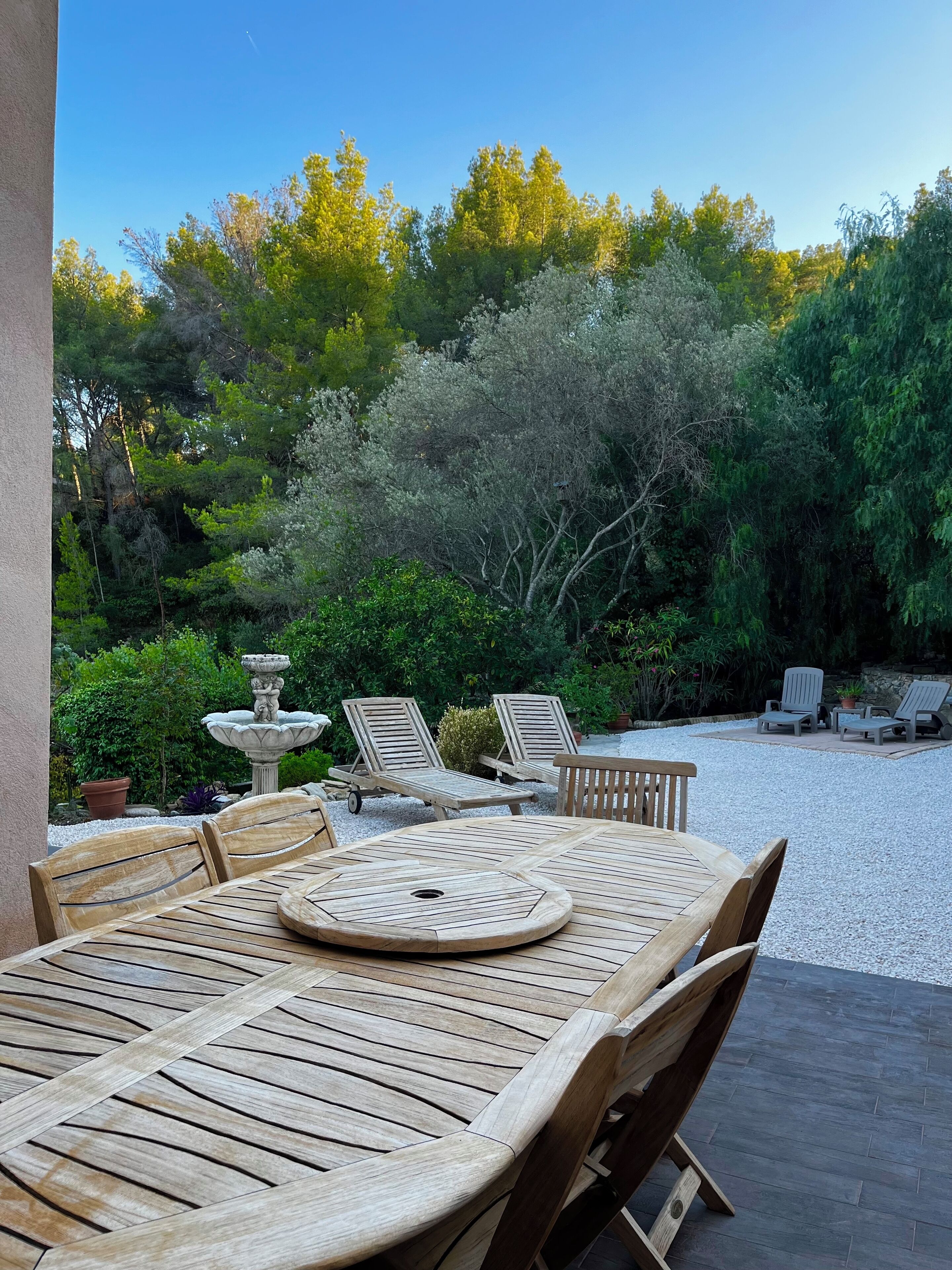 Un patio exterior con una mesa y sillas de madera, rodeado de grava y vegetación.