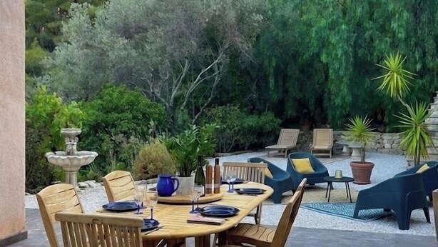 Een terras buiten met een houten tafel en stoelen, omgeven door groen en een stenen muur.