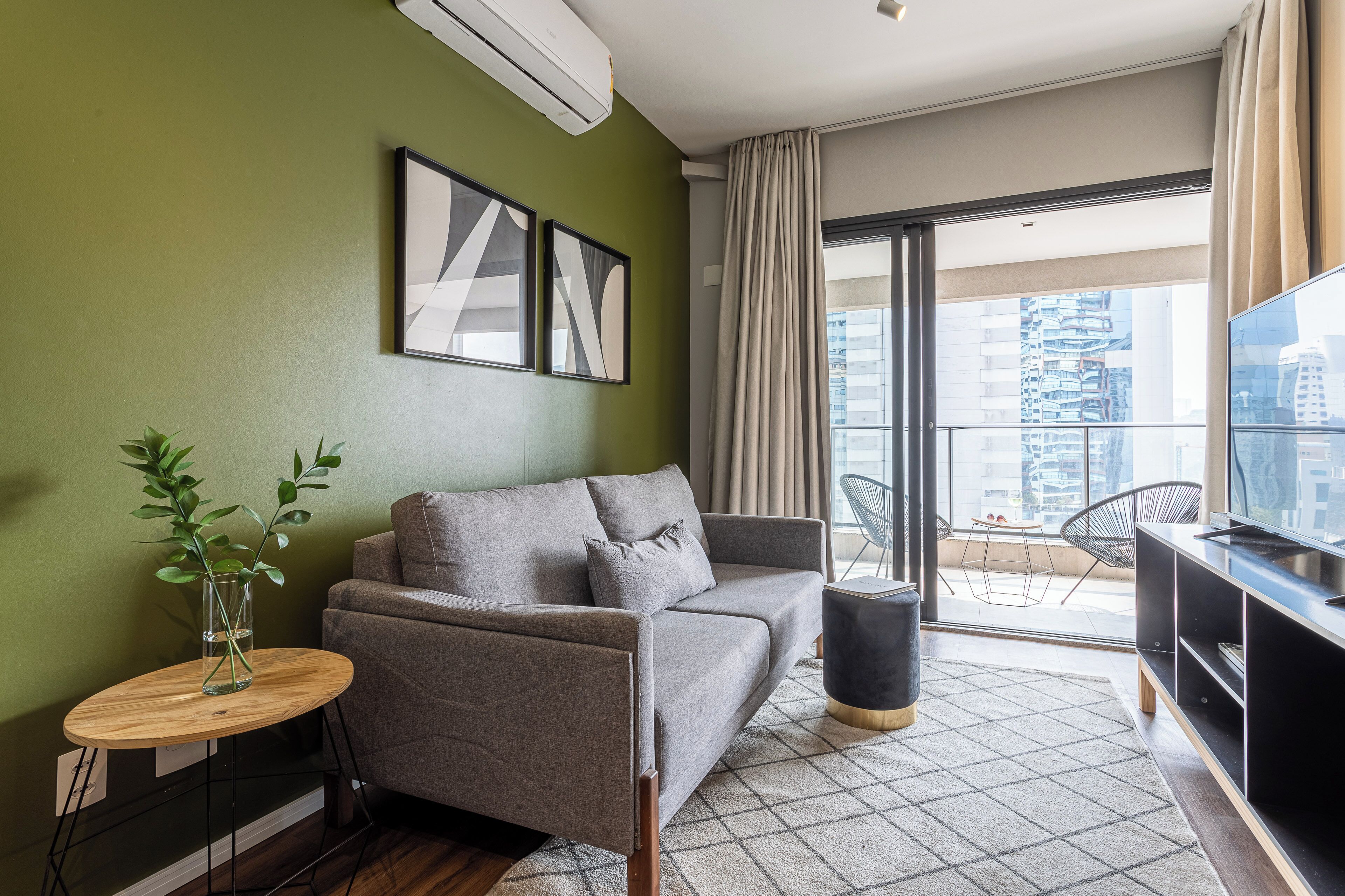 Una sala moderna con un sofá gris, una mesita redonda con una planta, un mueble de televisión negro y un ventanal con vistas a edificios.