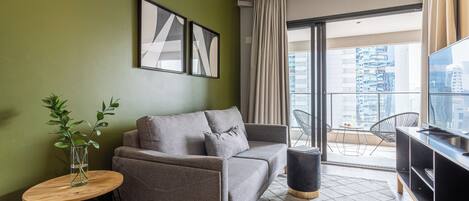 Una sala moderna con un sofá gris, una mesita redonda con una planta, un mueble de televisión negro y un ventanal con vistas a edificios.