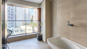 A modern bathroom with a bathtub, a potted plant, and a view of tall buildings through a large window.