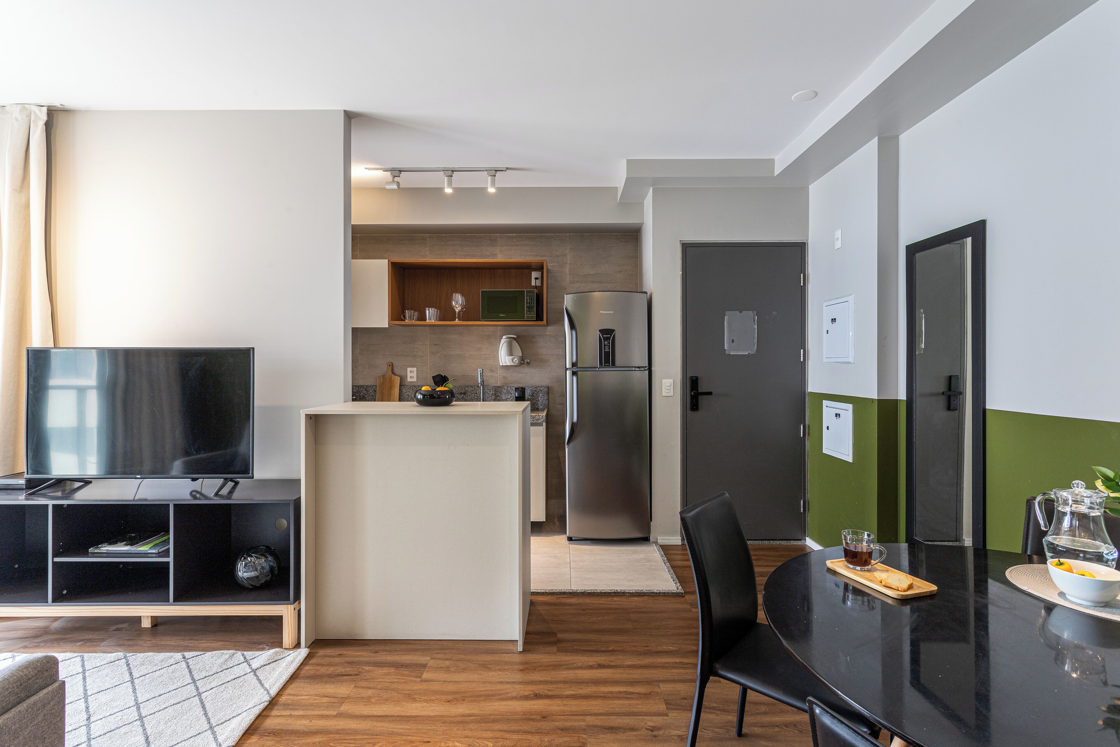 A modern kitchen with a built-in microwave, refrigerator, and a small island. There is a dining table with chairs and a TV in the living area.
