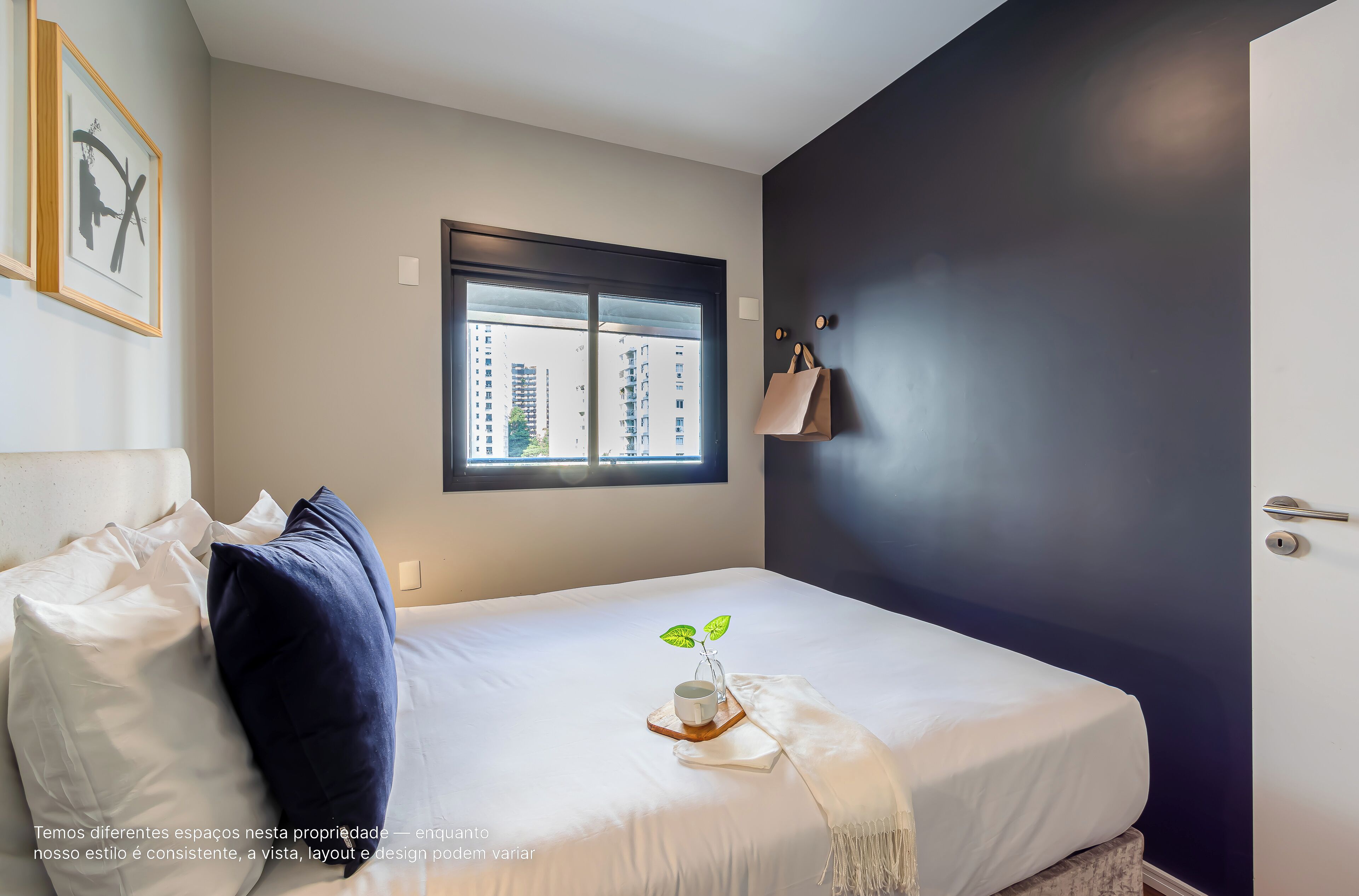 A neatly made bed with pillows, a bedside table with a cup and a small plant, and a window with a view of buildings.
