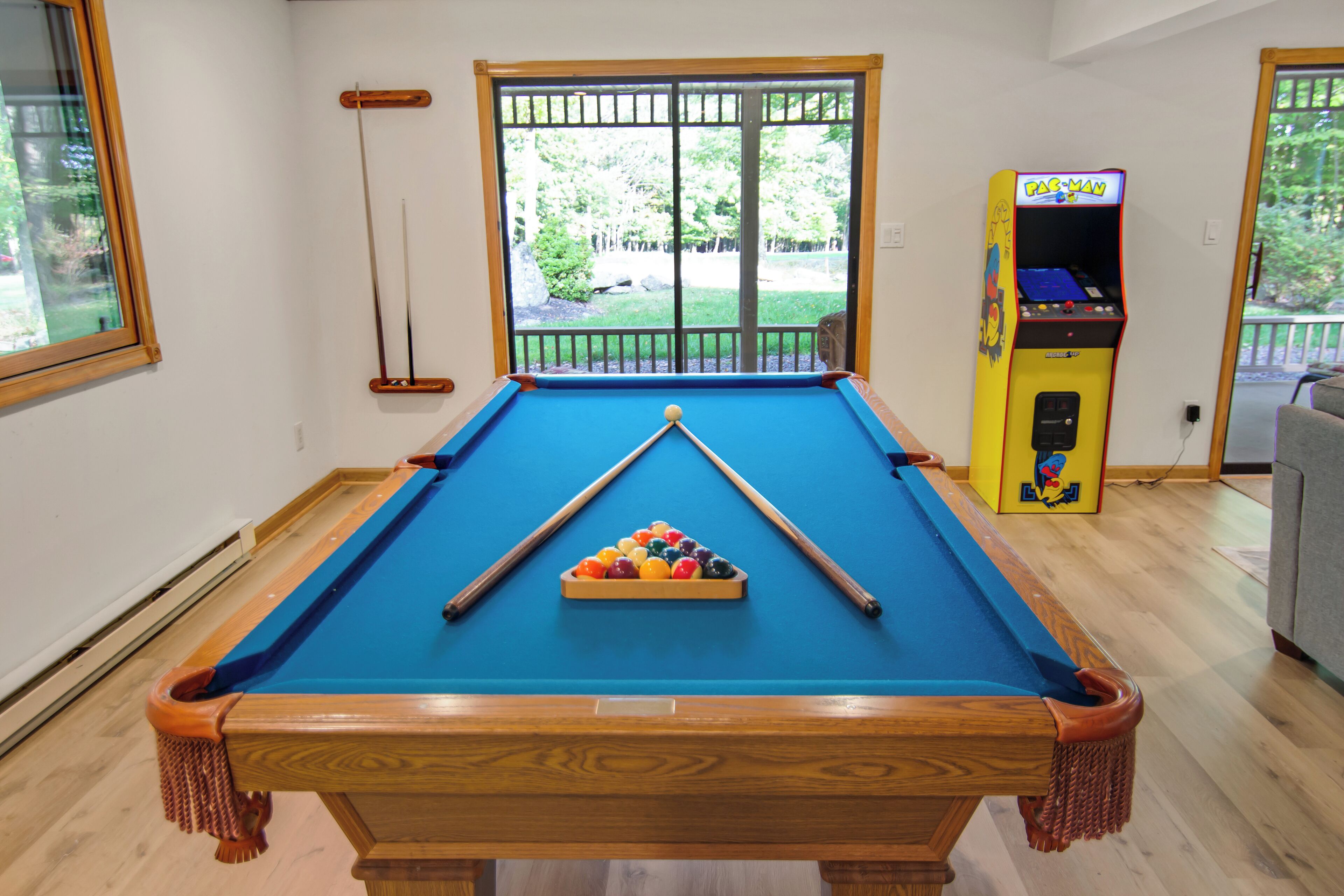 A pool table with a set of balls, a pool cue, and a Pac-Man arcade machine in a room with wooden flooring and a large window.