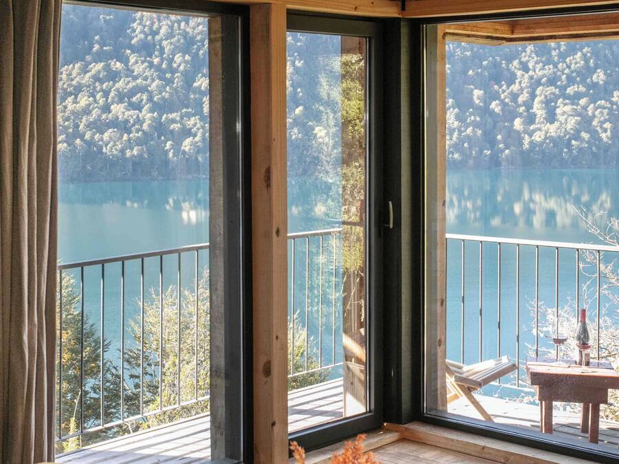 A room with a wooden ceiling, large windows, a balcony with a view of a lake, and two beds with towels.