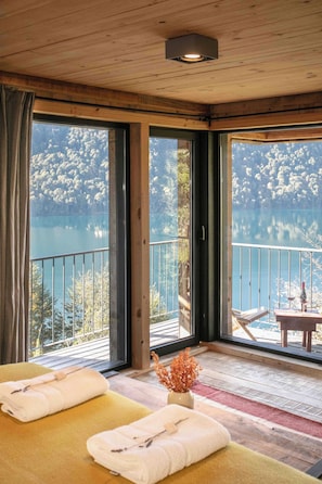 A room with a wooden ceiling, large windows, a balcony with a view of a lake, and two beds with towels. - Hermoso Eco Lodge (San Martín de los Andes)