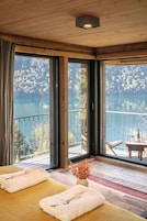 Quarto com teto de madeira, grandes janelas, varanda com vista para um lago e duas camas com toalhas.