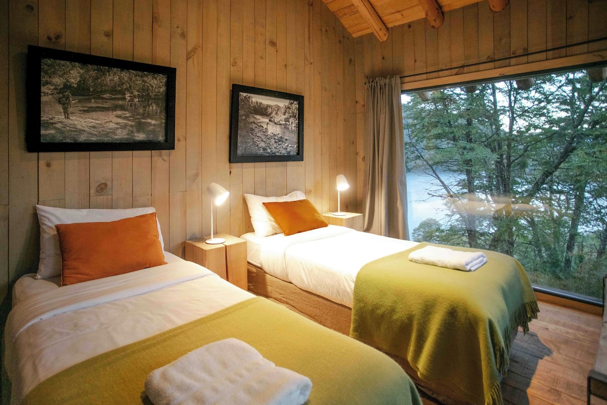 A wooden cabin room with two beds, framed pictures on the wall, and a window with a view of trees.