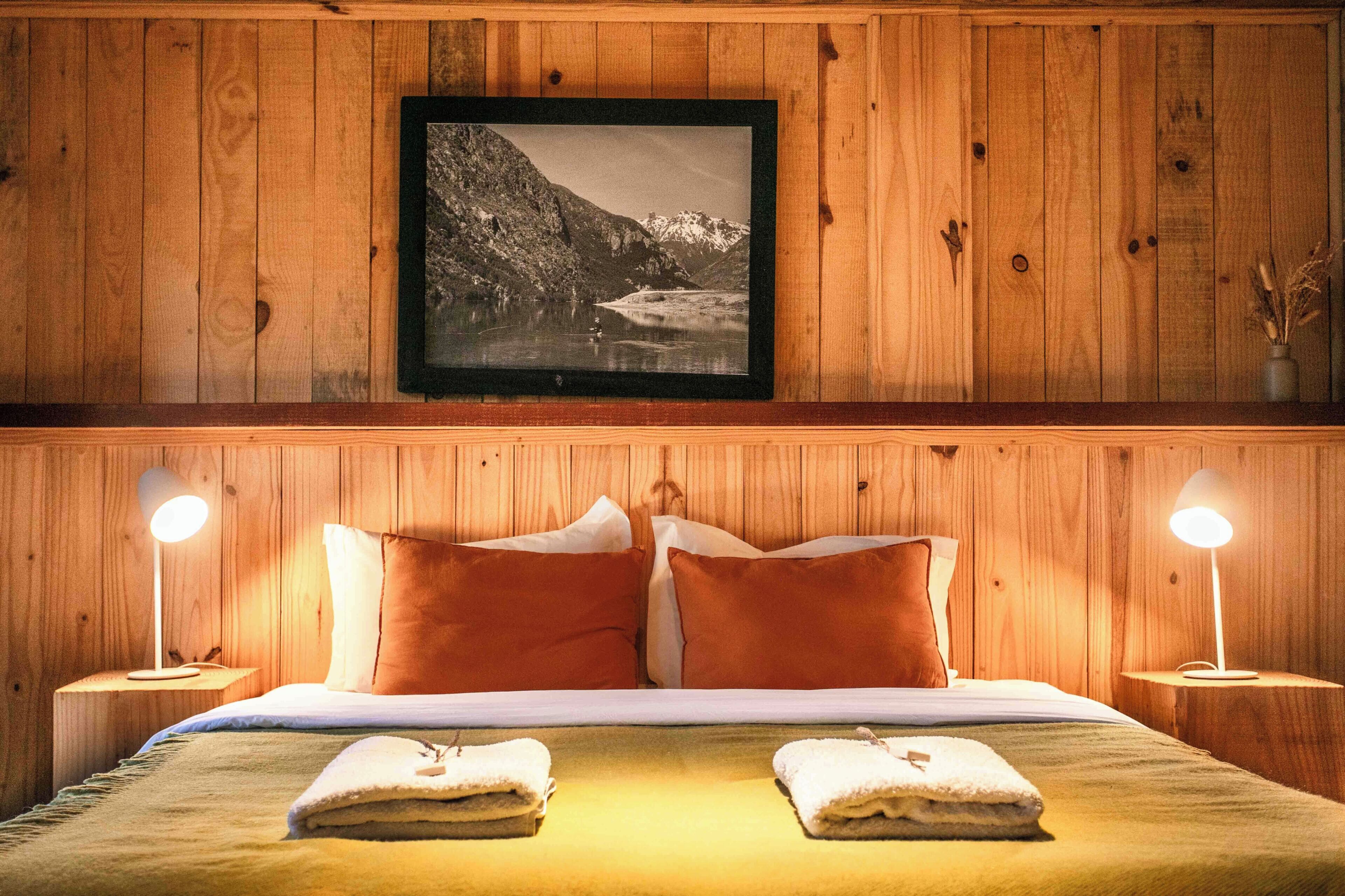 Een kamer met houten lambrisering, een bed, twee bedlampjes, een lijst met een afbeelding aan de muur en twee handdoeken op het bed.
