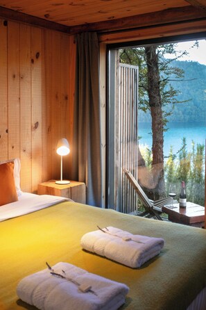 A wooden room with a bed, a nightstand with a lamp, a table with a glass of wine, and a view of a lake. - Hermoso Eco Lodge (San Martín de los Andes)