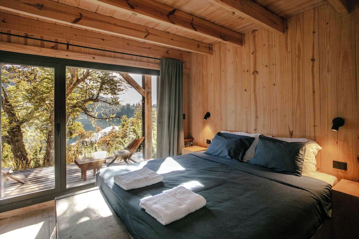 A wooden cabin bedroom with a large bed, two pillows, a nightstand, and a view of trees and mountains through large windows.
