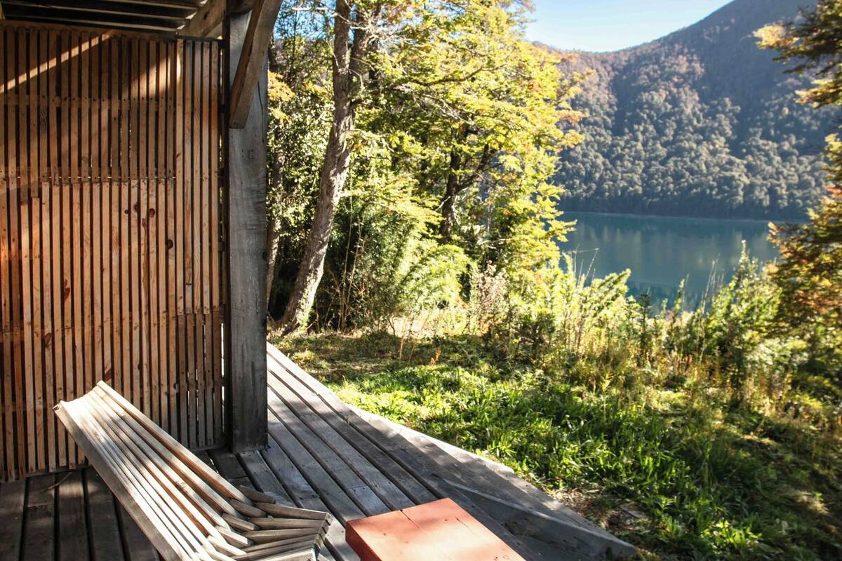A wooden deck with a bench and a small table, overlooking a lake and forested hills.