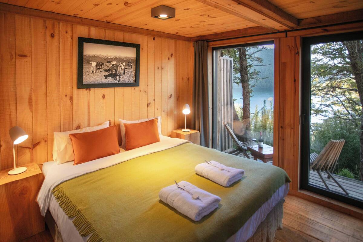 A wooden cabin bedroom with a bed, two bedside lamps, a framed photo, and a view of a lake.