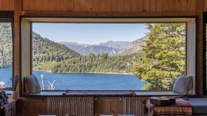 A room with a large window overlooking a lake and mountains, a wooden table with books and a guitar, and a cozy seating area with cushions. - Hermoso Eco Lodge (San Martín de los Andes)