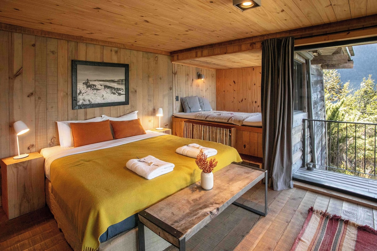 A wooden cabin-style bedroom with a bed, bedside lamps, a bench, a window with curtains, and a framed picture on the wall.